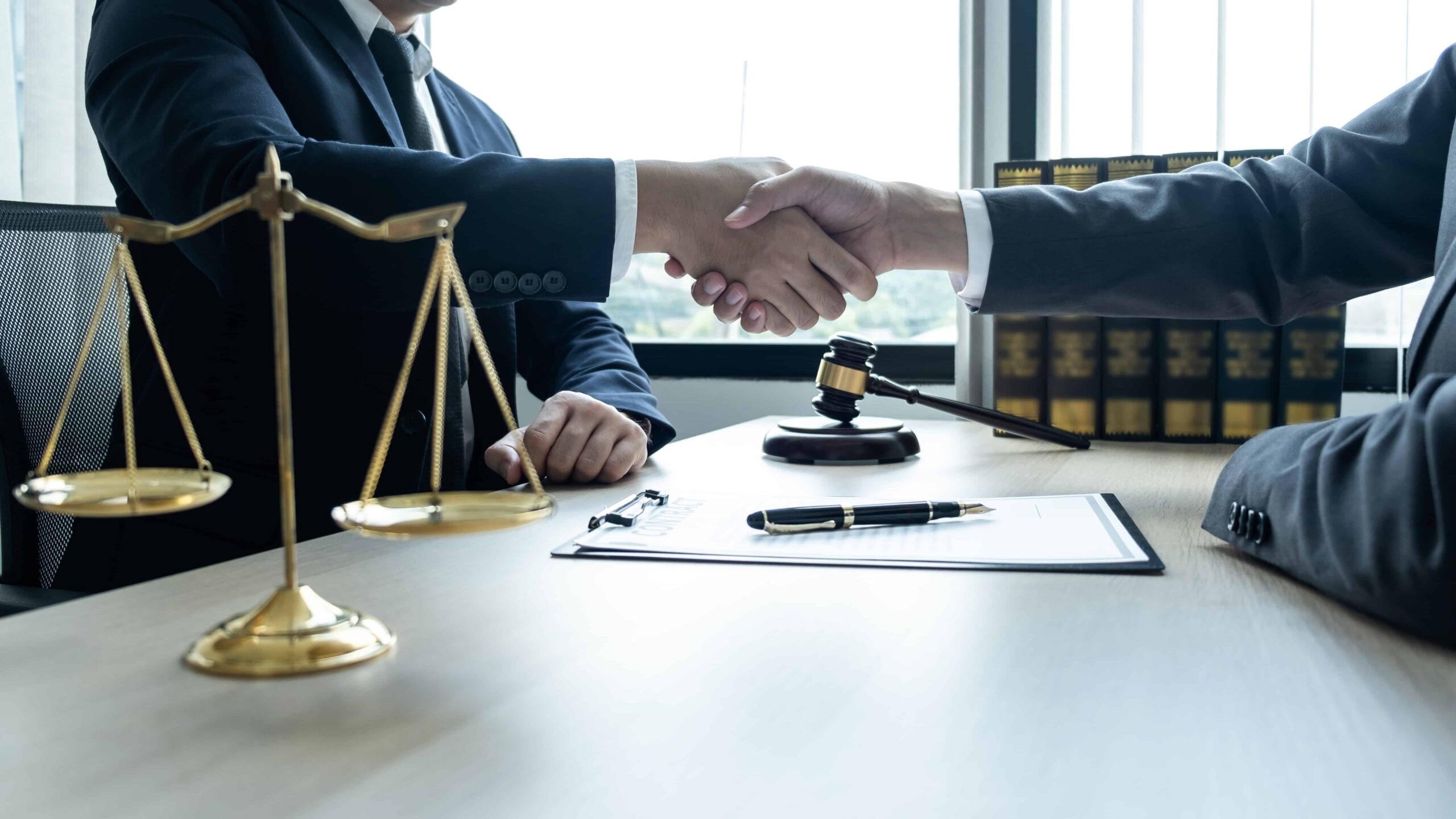 Lawyers shaking hands across a desk with a judge’s gavel and scales of justice, symbolizing a legal agreement or settlement.